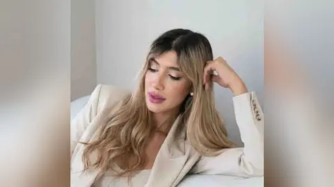 Blonde woman in a beige blazer posing thoughtfully in a bright, minimalist indoor setting.