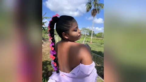 Young woman with long dark braid and pink flower accessories wearing a lavender top in a sunny tropical field.