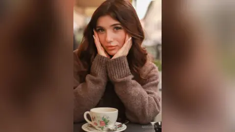 Young woman in a cozy brown sweater sitting at an outdoor cafe with a floral tea cup, looking thoughtful.