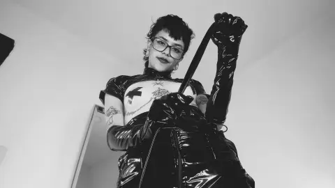 Edgy woman in black latex outfit and long gloves posing in a minimalist indoor setting, black and white aesthetic.