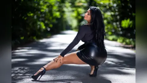 MissyJolie poses on a paved path wearing a black long-sleeve top and a tight black leather mini skirt with high heels.