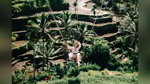 StacyHarland sits on a wooden swing suspended by long ropes over a lush, terraced tropical jungle landscape.