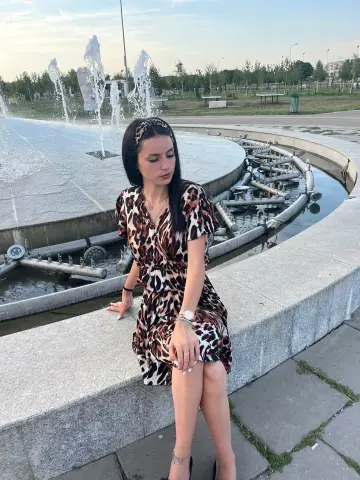 Young woman in a leopard print dress sitting by a stone fountain in a peaceful park at sunset.