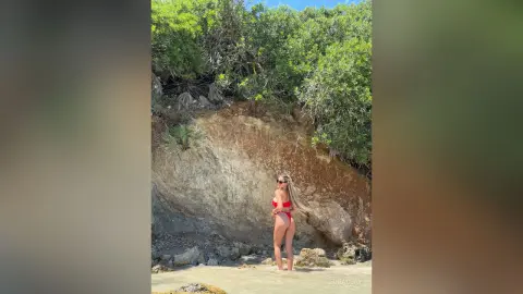 Woman in a red bikini standing by a rocky cliff under lush green trees on a sunny summer day.