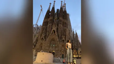 LucyMartin stands in front of the Sagrada Familia, wearing a white tank top and light blue denim jeans.