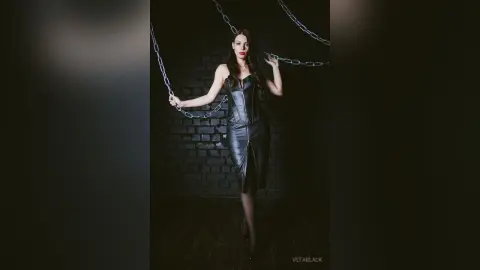 Elegant woman in a black leather corset dress posing against dark brick walls with hanging metal chains.