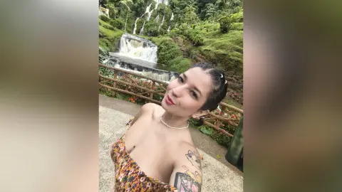 Smiling young woman in a floral sundress relaxing by a lush tropical waterfall in a serene jungle setting.