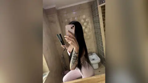 AriaVex takes a mirror selfie in a bathroom, showing her long black hair and arm tattoos while wearing light pink leggings.