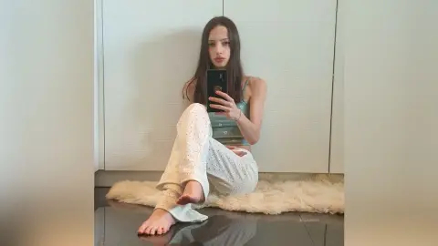 Young woman in white patterned lounge pants and green tank top sitting on a fluffy rug in a minimalist room.