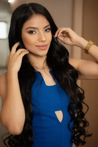 Young woman with long wavy black hair wearing a blue sleeveless dress and gold necklace in a soft indoor setting.