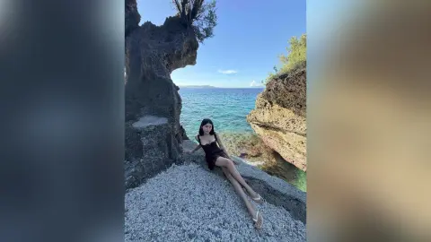 Young woman in a black summer dress relaxing on rocky cliffs overlooking a turquoise tropical ocean.
