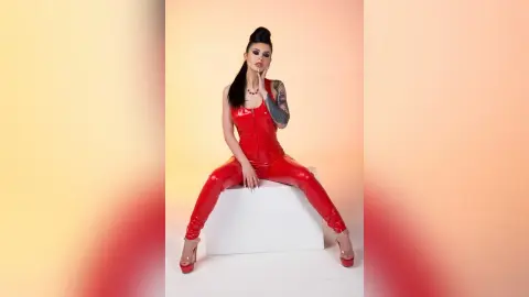 Confident woman in a red shiny jumpsuit posing on a white pedestal against a warm studio background.