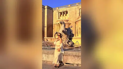 Stylish woman in a white summer outfit posing by a classical stone fountain in warm golden hour sunlight.