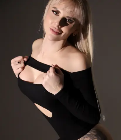 Blonde woman in a black off-the-shoulder bodysuit posing against a dark studio background.