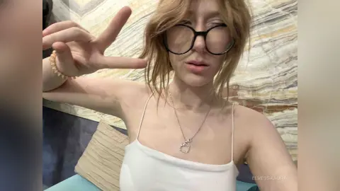 Blonde woman in a white camisole and glasses making a peace sign in a cozy indoor setting.