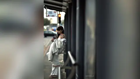 Stylish woman in white outfit waiting at a city bus stop on a moody urban afternoon.
