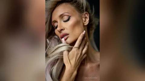 LeslyAnderson poses with her eyes closed, resting her hand against her face and neck while showing her blonde hair and makeup.