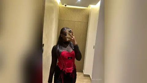 AmyJacobs poses in a mirror wearing a red corset top and black long-sleeve mesh shirt, standing in a hallway with tan slatted walls.