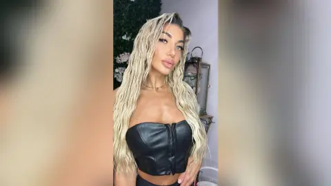 Blonde woman in a black leather corset top posing with a confident expression in a modern indoor setting.