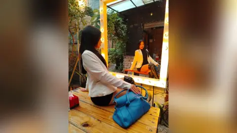 EvieMillers sits on a wooden bench wearing a white blazer and black shorts, looking into a bright vanity mirror with a blue handbag nearby.