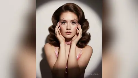 TeresiaCaspi poses with her hands on her face, wearing pink makeup gems and a pink strap against a plain background.