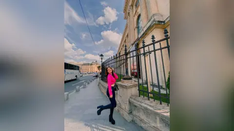 Smiling woman in a pink top and black trousers standing by a black metal fence on a sunny city street.
