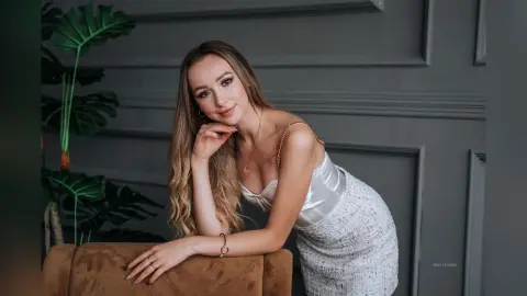 Smiling young woman in a white satin dress posing elegantly against a grey paneled wall in a modern interior.