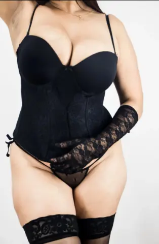 Curvy woman in elegant black lace lingerie, corset, and sheer gloves posing against a white background.