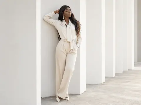 Elegant Black woman in cream silk blouse and trousers leaning against white pillars in a bright, minimalist setting.