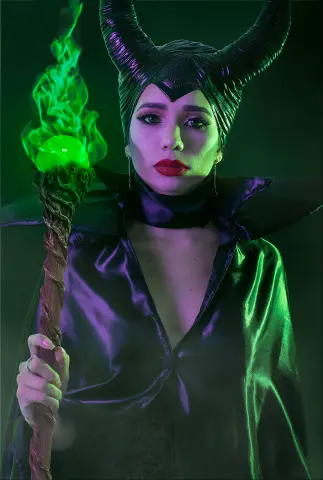 Woman in Maleficent cosplay holding a glowing green magic staff in a dark, mystical fantasy setting.