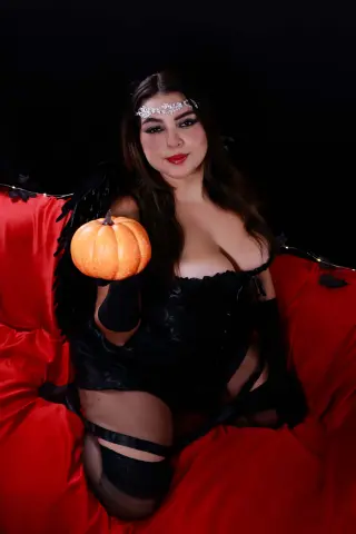 Woman in black gothic lingerie and crown sitting on a red velvet sofa holding a pumpkin for Halloween.