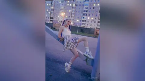 Young woman in a white tank top and plaid skirt sitting on a ledge against a city building at dusk.