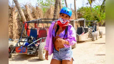 Adventurous woman in a purple shirt and blue helmet standing by off-road buggies on a sunny tropical trail.