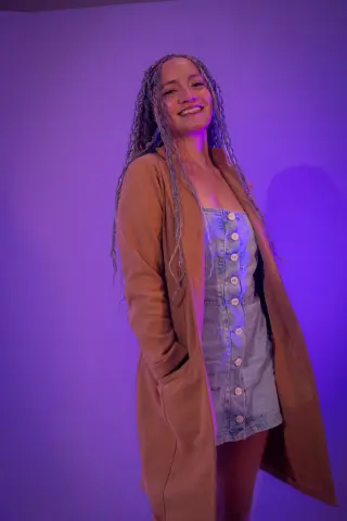 Smiling woman with long braids wearing a denim dress and brown cardigan in moody purple studio lighting.