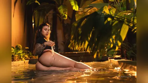 MilaWillow sits in a pool surrounded by tropical plants, wearing a black bikini and looking back over her shoulder.