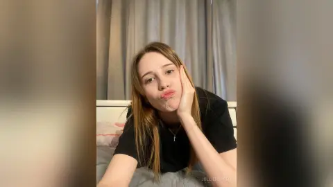 Young woman with long blonde hair in a black t-shirt making a playful pouty face in a cozy bedroom setting.