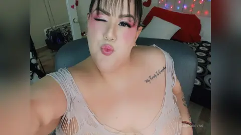 Woman with pink makeup and white lace top making a playful kissy face in a cozy bedroom setting.
