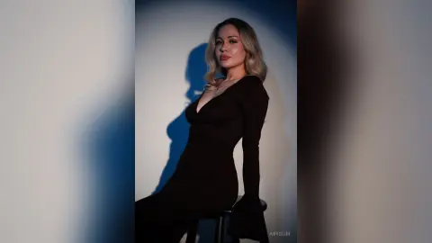 Elegant blonde woman in a chic black long-sleeve dress posing against a blue studio background with dramatic lighting.