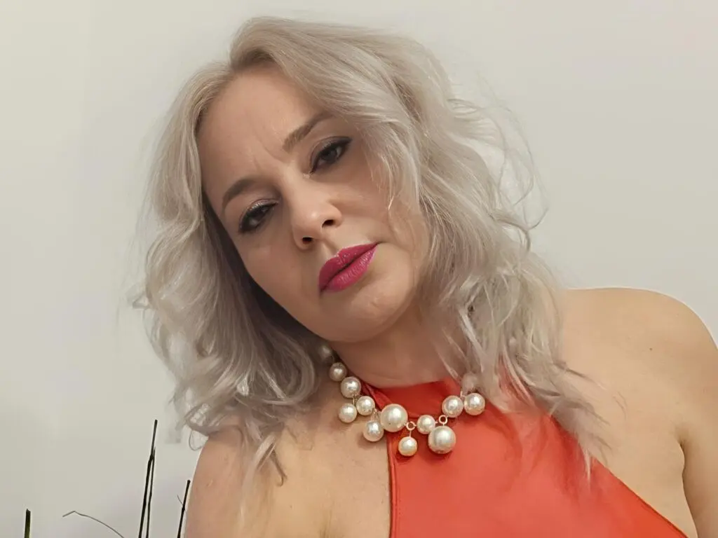 Blonde woman in a bright orange halter top and chunky pearl necklace posing against a white background.