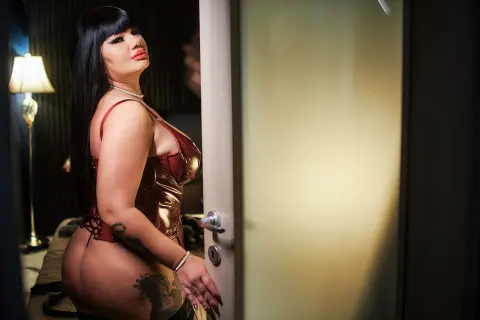 Curvy woman in a red metallic bodysuit posing in a dimly lit bedroom setting.