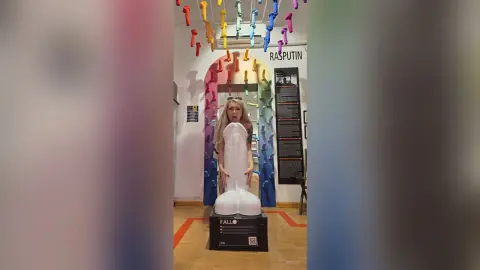 Blonde woman in a black top standing in a quirky room decorated with colorful hanging phallic art decor.