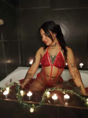 Tattooed woman in a red sequin bikini relaxing in a bathtub surrounded by glowing candles in a moody setting.