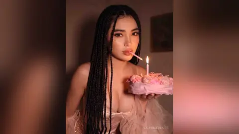 Young woman with long braids holding a pink birthday cake with a lit candle in a soft, moody indoor setting.