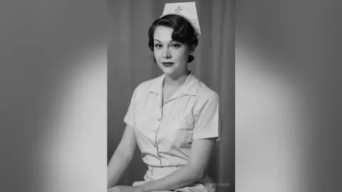 Vintage portrait of a smiling nurse in a classic white uniform and cap against a neutral studio background.