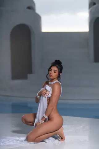 Glamorous woman in a white draped outfit posing by a luxury white Mediterranean villa pool at dusk.