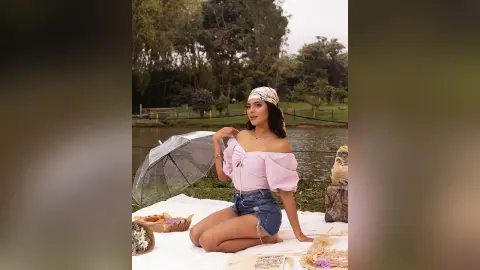 Young woman in a pink off-the-shoulder top and denim shorts relaxing on a picnic blanket by a serene lake.