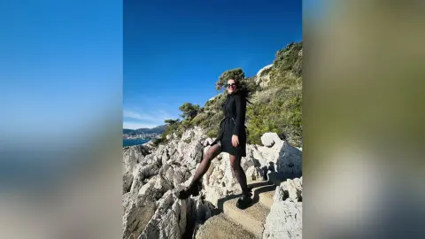 VesperMuse poses outdoors on a rocky path wearing a short black dress, black tights, and sunglasses against a clear blue sky.