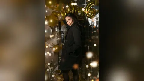 Elegant woman in a black coat posing amidst gold balloons at a festive celebratory party.