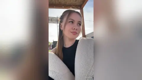Young woman in a black top and cozy white fleece jacket looking away under a rustic wooden patio canopy.