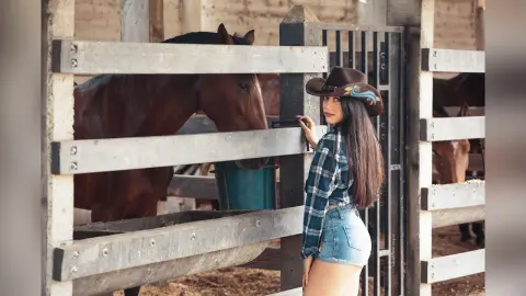 JessieLiu stands next to a wooden fence in a stable, wearing a blue plaid shirt and denim shorts.